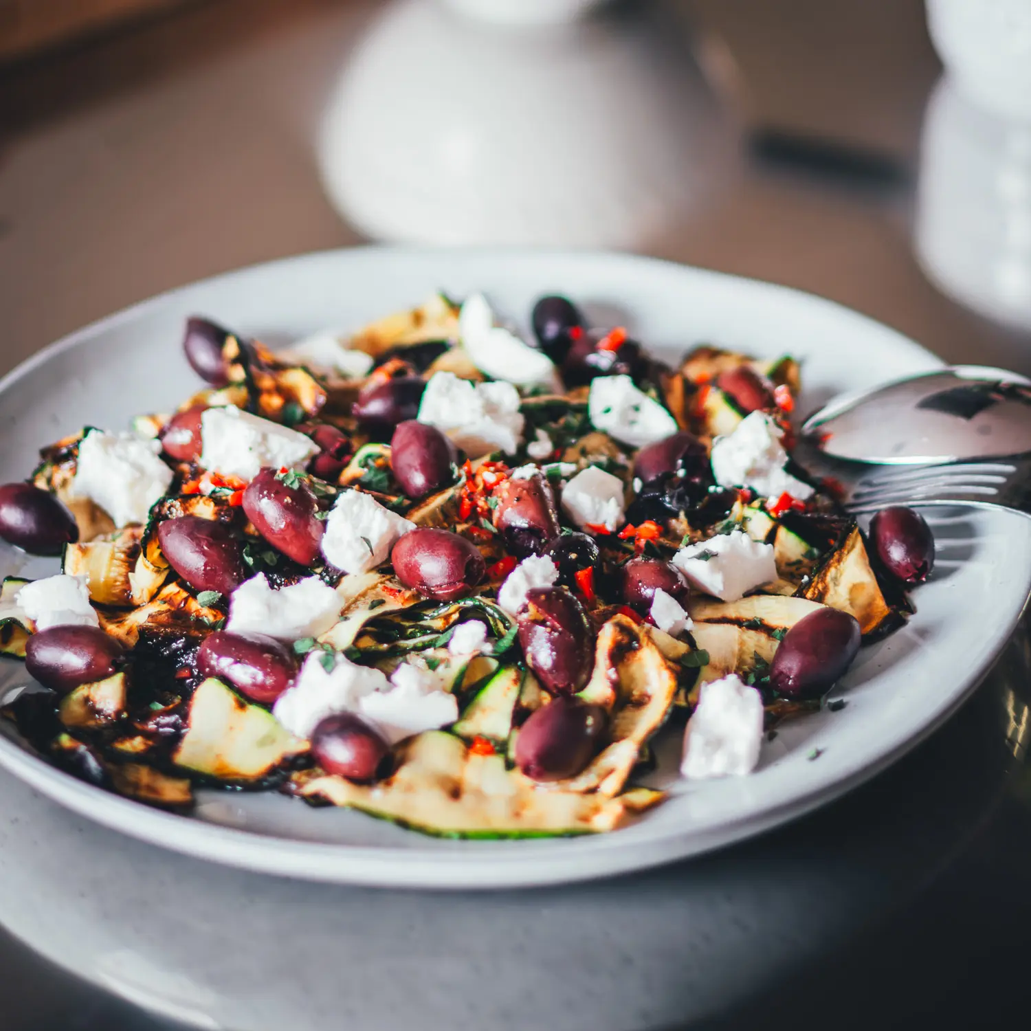 Angerichteter gegrillter Zucchini-Salat mit Oliven und Ziegenfrischkäse auf hellem Teller, im Hintergrund die Küche