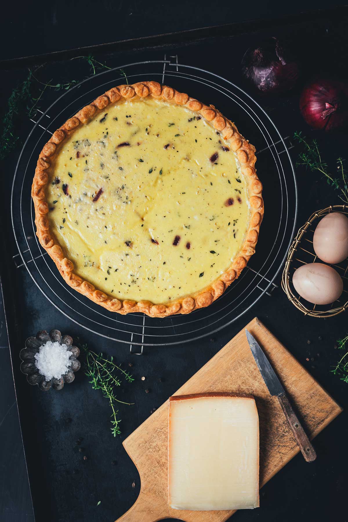Zwiebelquiche von oben fotografiert, ganze Quiche mit karamellisierten roten Zwiebeln, daneben Thymian, Eier, Bergkäse, Salz und Zwiebeln als Deko | moeyskitchen.com