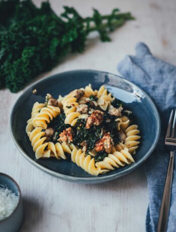 Dunkelblaue Schale mit Pastagericht mit kurzen Nudeln, Grünkohl und Bratwurst auf hellem Holz. Im Hintergrund mit Grünkohl und einer hellblauen Serviette.