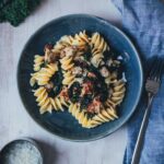 Topshotaufnahme eines dunkelblauen Tellers, gefüllt mit Pasta mit Grünkohl und Bratwurstbrät und Senfsauce. Daneben liegt eine kleine Schüssel mit geriebenem Parmesan, einige Blätter Grünkohl und eine hellblaue Serviette mit einem Esslöffel