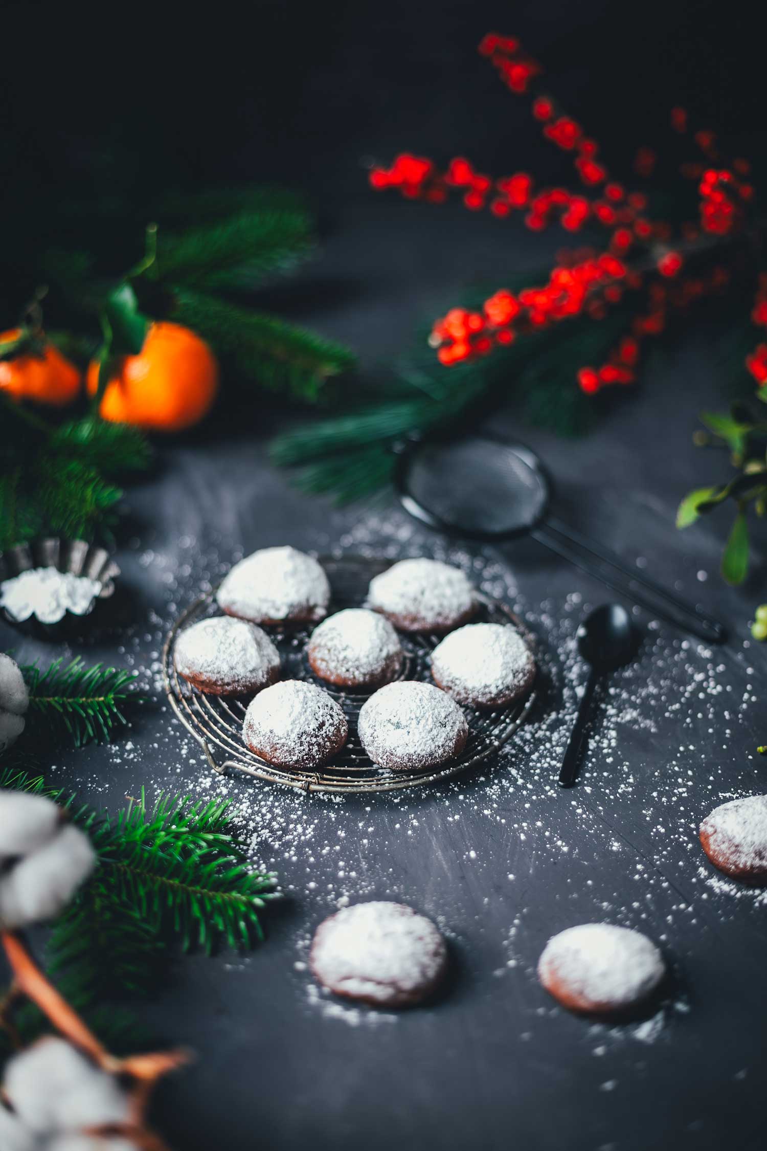 Mürbe Espresso-Walnuss-Kekse auf einem Gitter, mit Puderzucker bestäubt und weihnachtlicher Deko aus Tannengrün und Orangen im Hintergrund.