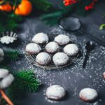 Seitliche Ansicht von mit Puderzucker bestreuten dunklen, runden Plätzchen auf einem kleinen Kuchengitter. Drumherum weihnachtliche Deko wie Tannenzweige, dazu ein Löffel und Sieb mit Puderzucker.