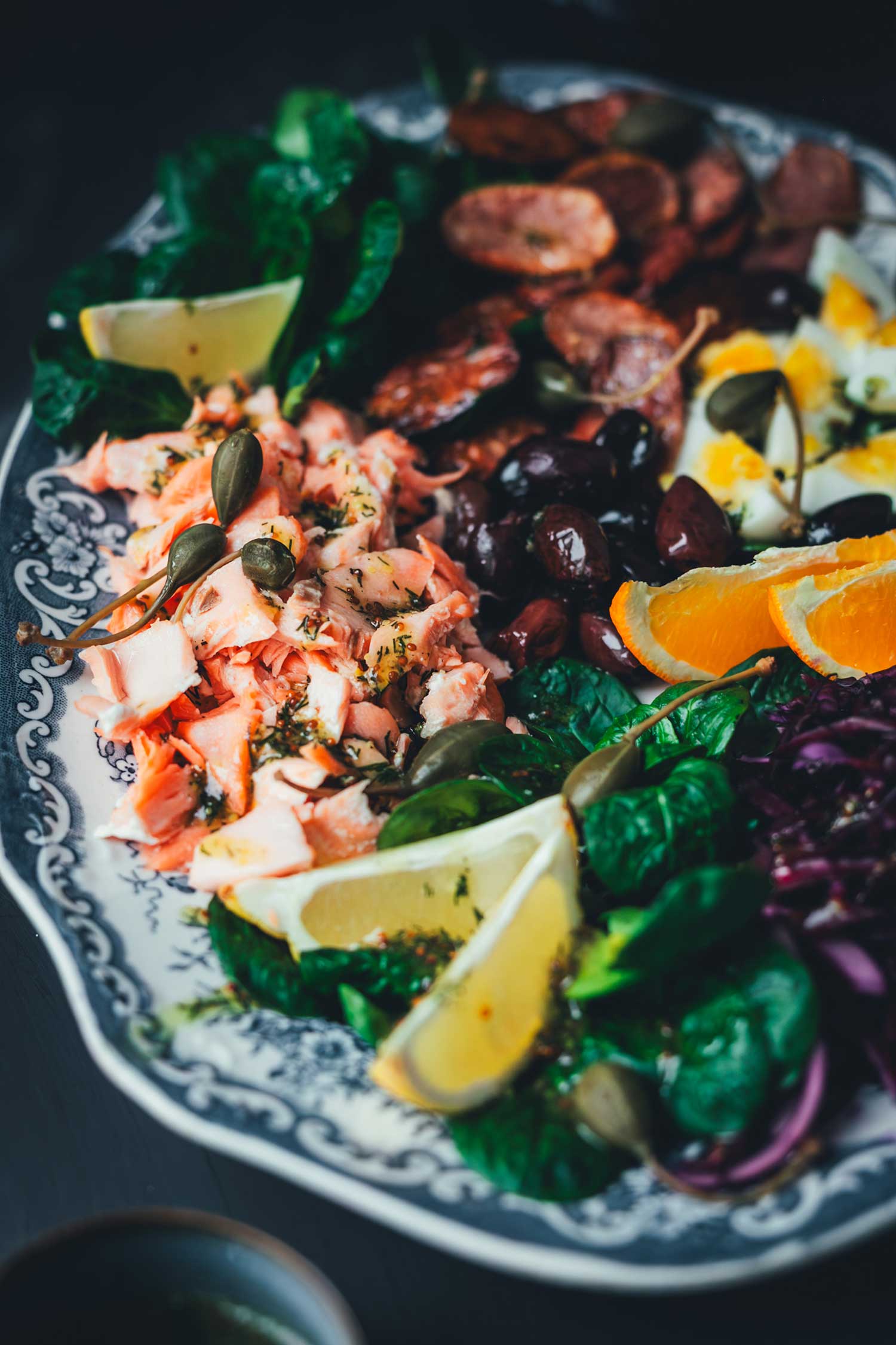 Nahaufnahme Winterlicher Nizza Salat mit Pulled Lachs auf Servierplatte – mit Rotkohl, Feldsalat, knusprigen Kartoffeln, Eiern, Orangen und Kapernäpfeln. Fokus auf das Lachsfilet.