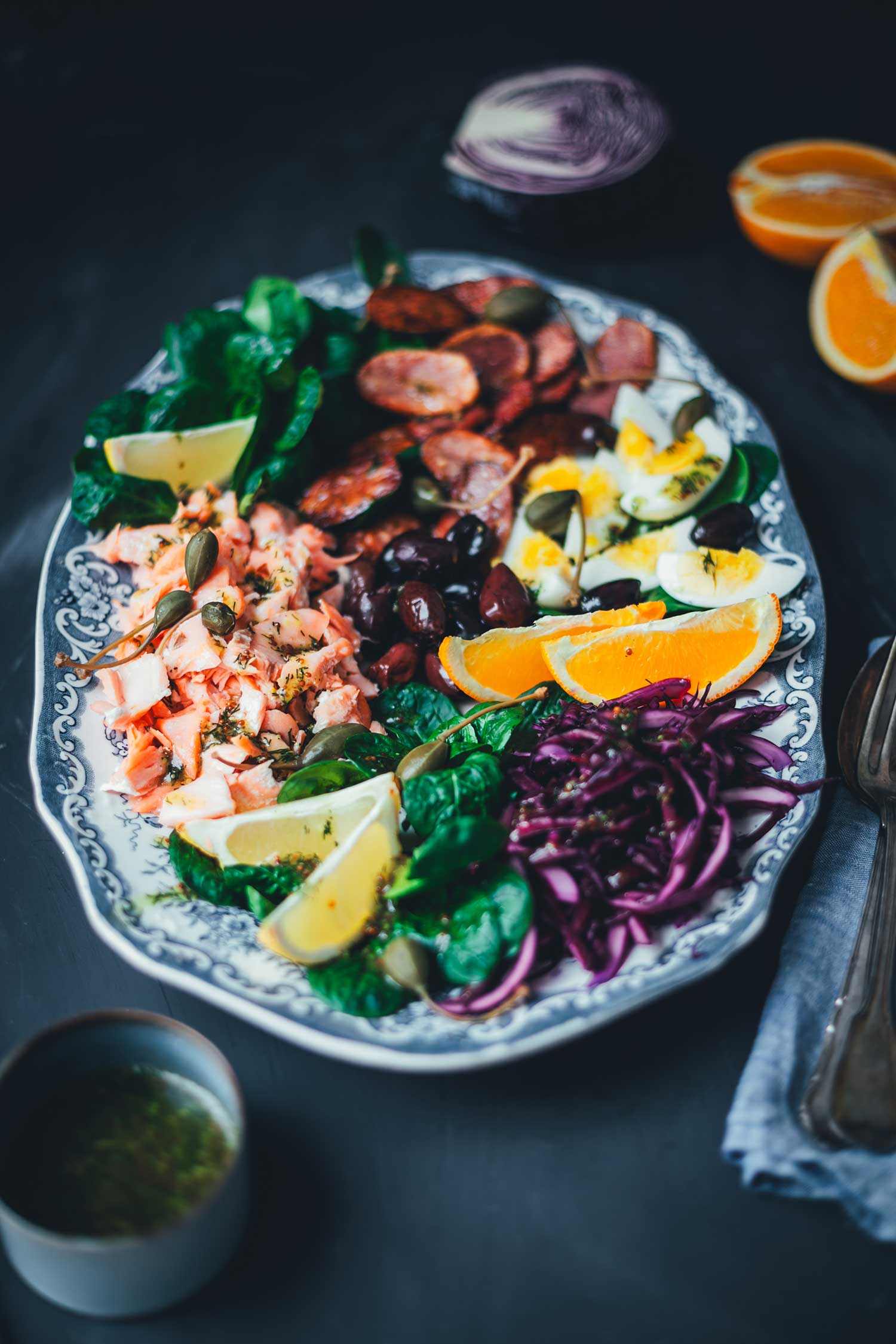 Seitliche Ansicht Winterlicher Nizza Salat mit Pulled Lachs auf Servierplatte – mit Rotkohl, Feldsalat, knusprigen Kartoffeln, Eiern, Orangen und Kapernäpfeln.