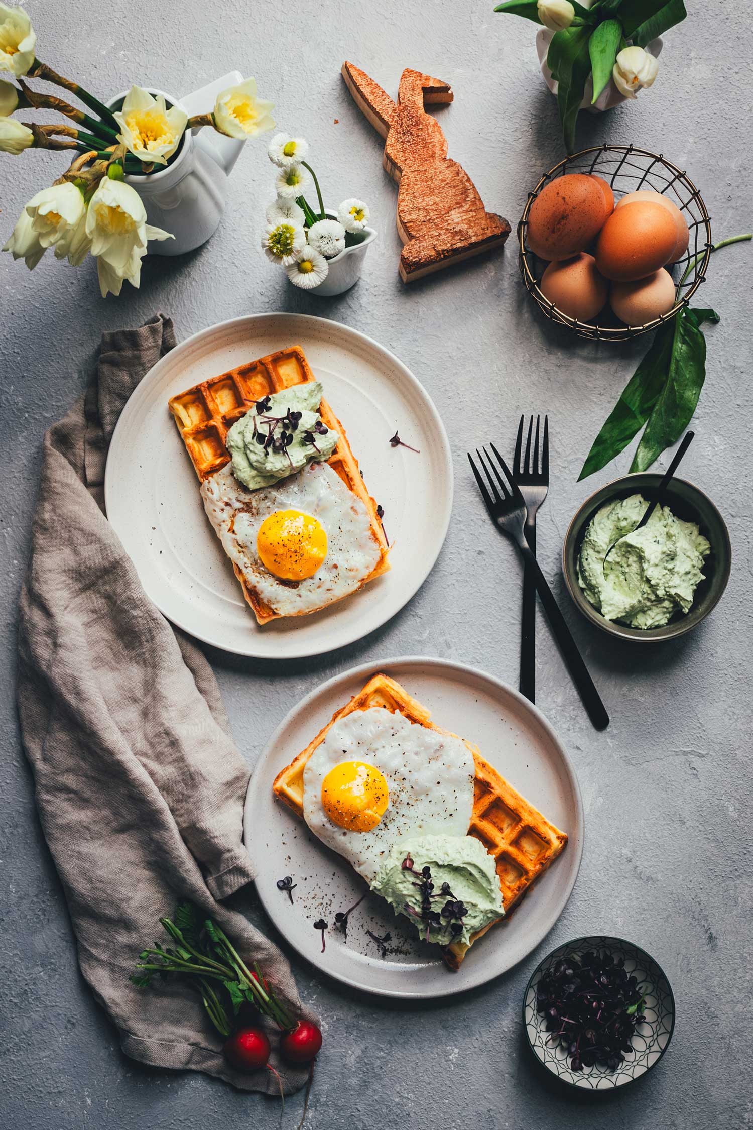 Topshot Aufnahme von einem gedeckten Tisch mit zwei servierfertigen Tellern mit frisch gebackenen herzhaften Kartoffel-Waffeln, getoppt mit Spiegelei und Bärlauch-Feta-Dip. Drumherum sieht man frische Kräuter und Gemüse, Frühlingsblumen und etwas österliche Deko.