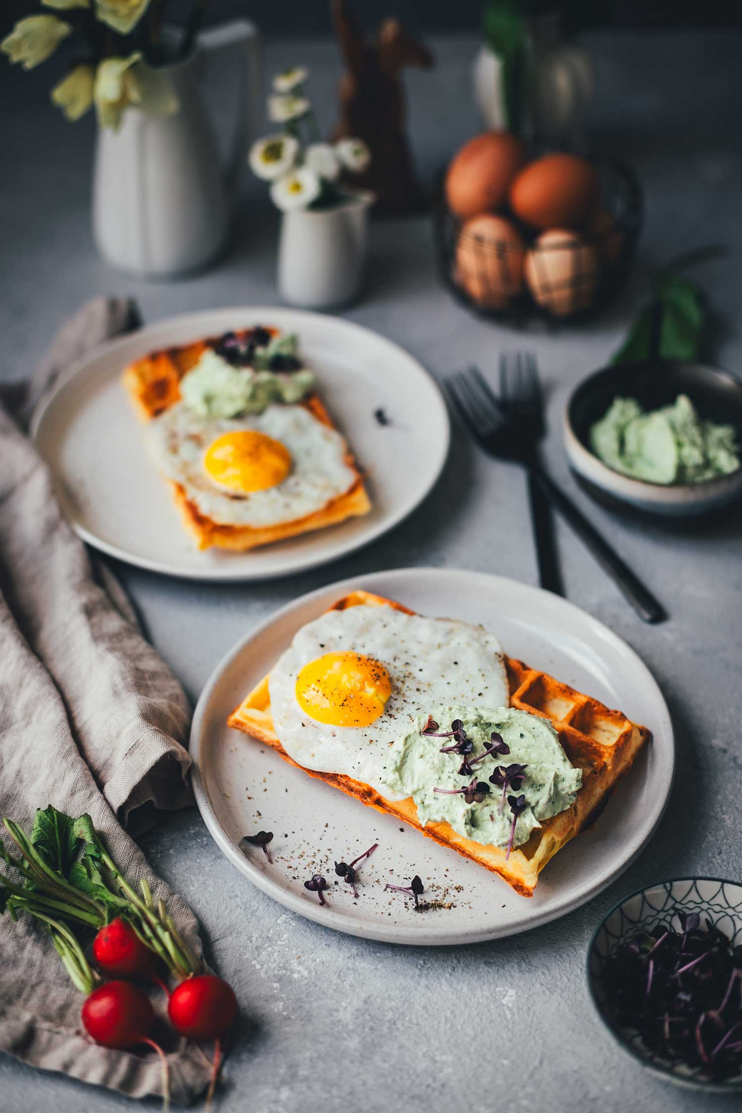 Leicht schräge Ansicht von einem gedeckten Tisch mit österlicher Deko sowie zwei Tellern mit frisch gebackenen herzhaften Waffeln mit Spiegelei und Bärlauch-Dip.