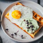 Nahaufnahme von einer herzhaften Kartoffel-Waffel auf einem Teller. Getoppt mit einem Spiegel, einer Bärlauch-Feta-Creme und Sprossen.