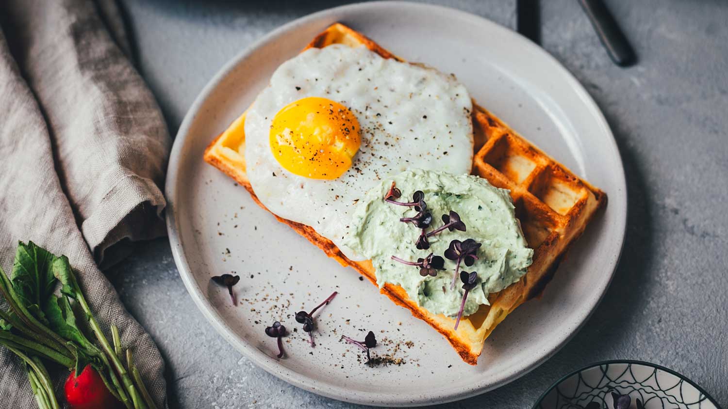 Nahaufnahme von einer herzhaften Kartoffel-Waffel auf einem Teller. Getoppt mit einem Spiegel, einer Bärlauch-Feta-Creme und Sprossen.
