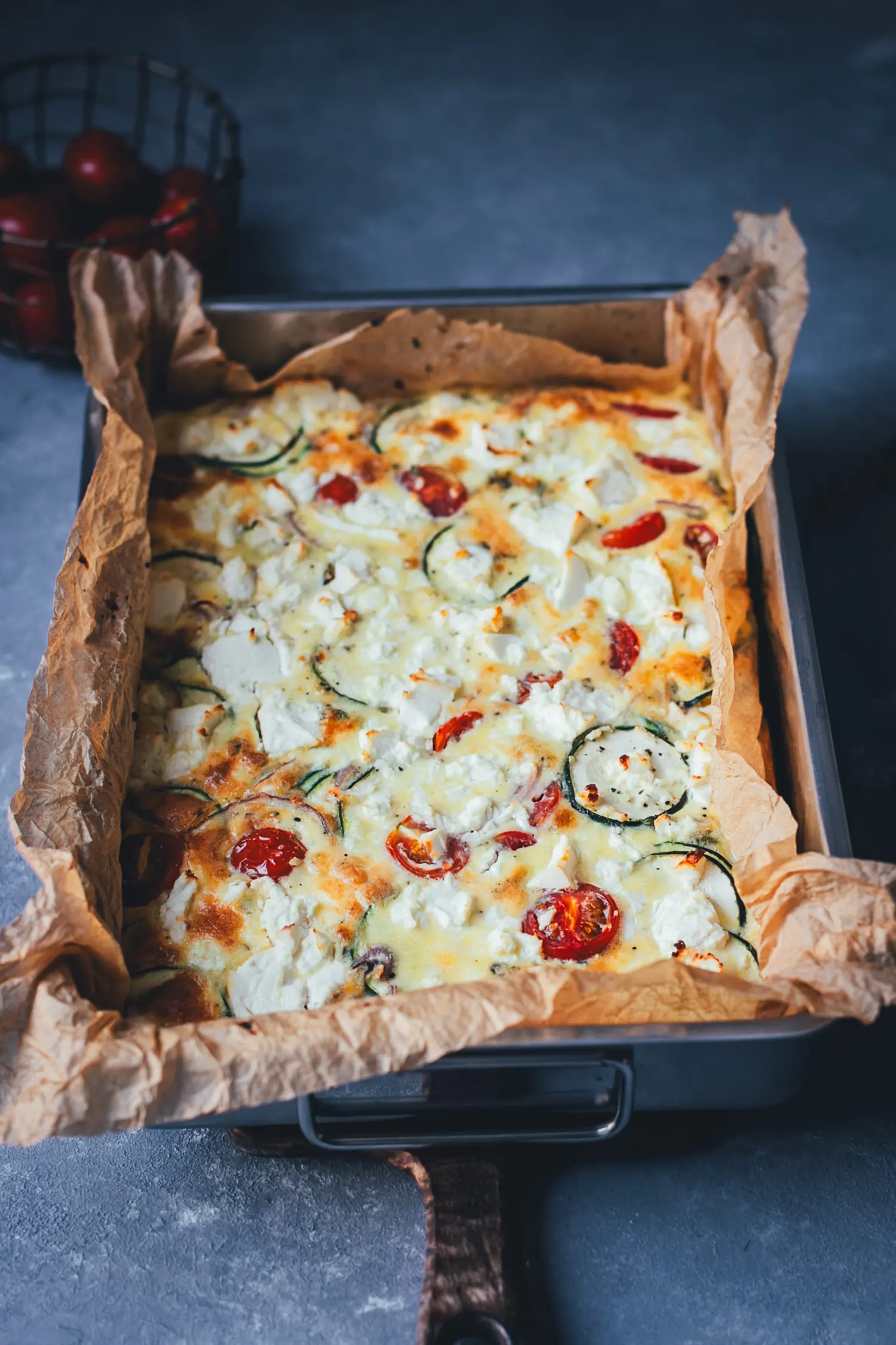 Goldbraun gebackene Zucchini-Frittata mit Feta und Tomaten in der Form, schräg von der Seite fotografiert.