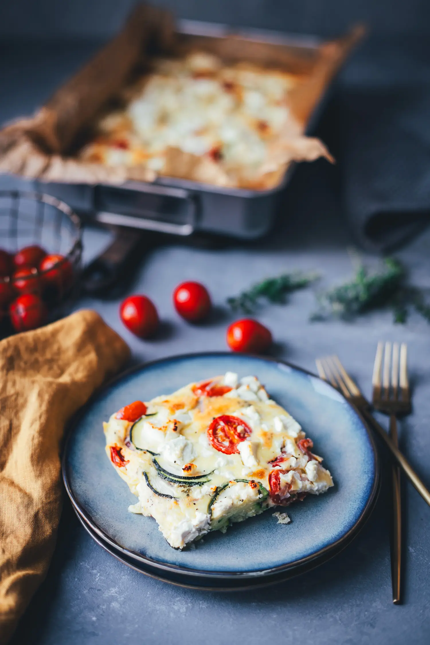 Ein Stück Zucchini-Frittata mit Feta und Tomaten auf einem blauen Teller, im Hintergrund die Backform.
