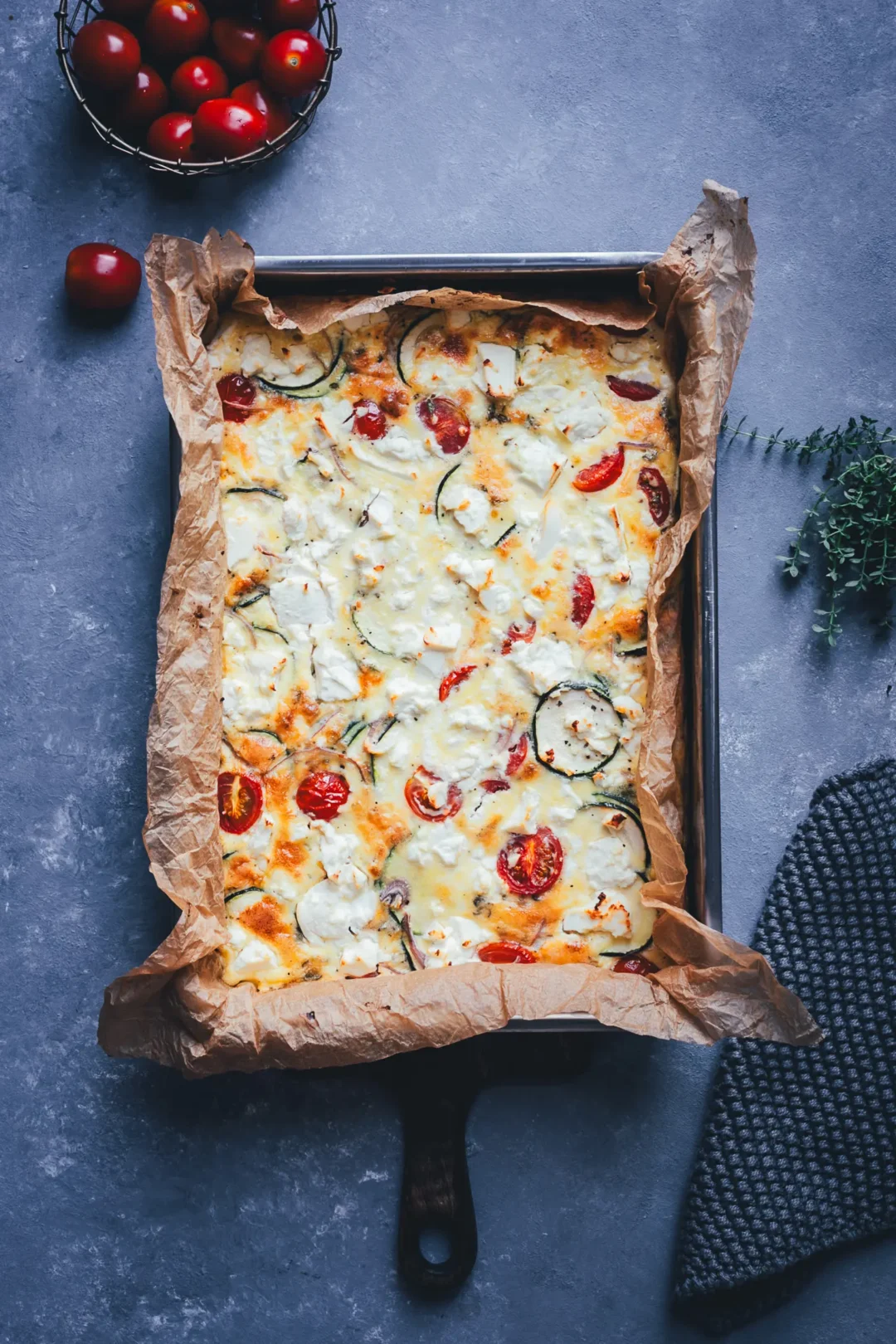 Zucchini-Frittata mit Feta und Tomaten aus dem Ofen, frisch gebacken in einer mit Backpapier ausgelegten Form, daneben Cherrytomaten und frischer Thymian.