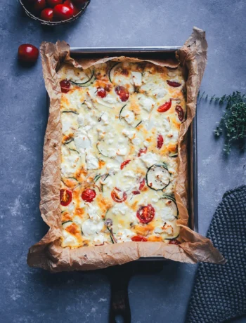 Zucchini-Frittata mit Feta und Tomaten aus dem Ofen, frisch gebacken in einer mit Backpapier ausgelegten Form, daneben Cherrytomaten und frischer Thymian.