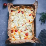 Quadratische Topshot-Aufnahme der fertigen Zucchini-Frittata mit Feta, Cherrytomaten und Zucchinischeiben in der Backform.