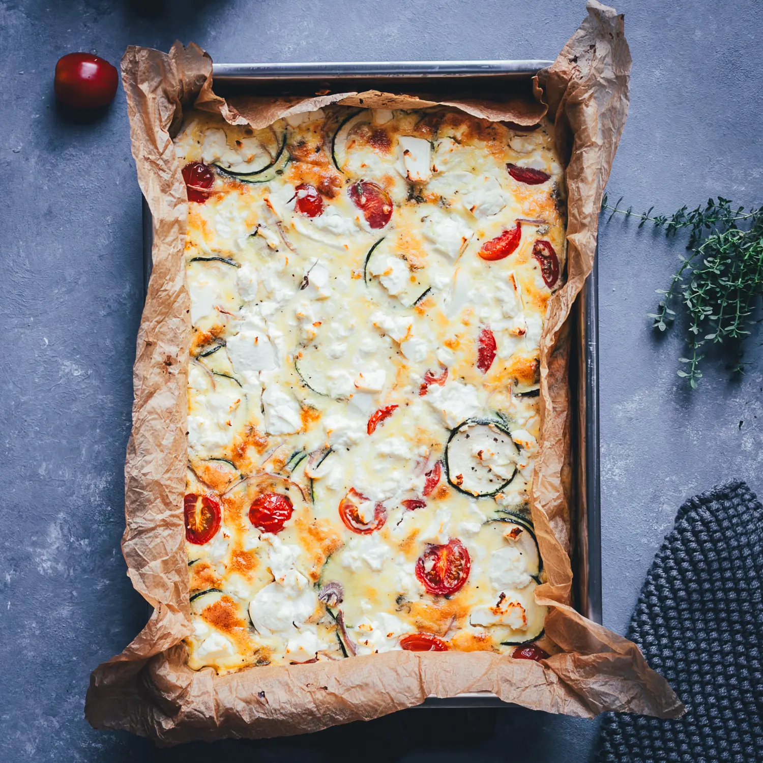 Quadratische Topshot-Aufnahme der fertigen Zucchini-Frittata mit Feta, Cherrytomaten und Zucchinischeiben in der Backform.