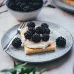 Nahaufnahme eines Stücks Hot Milk Sponge Cake mit Mascarpone-Creme und Brombeeren auf einem gesprenkelten Teller.