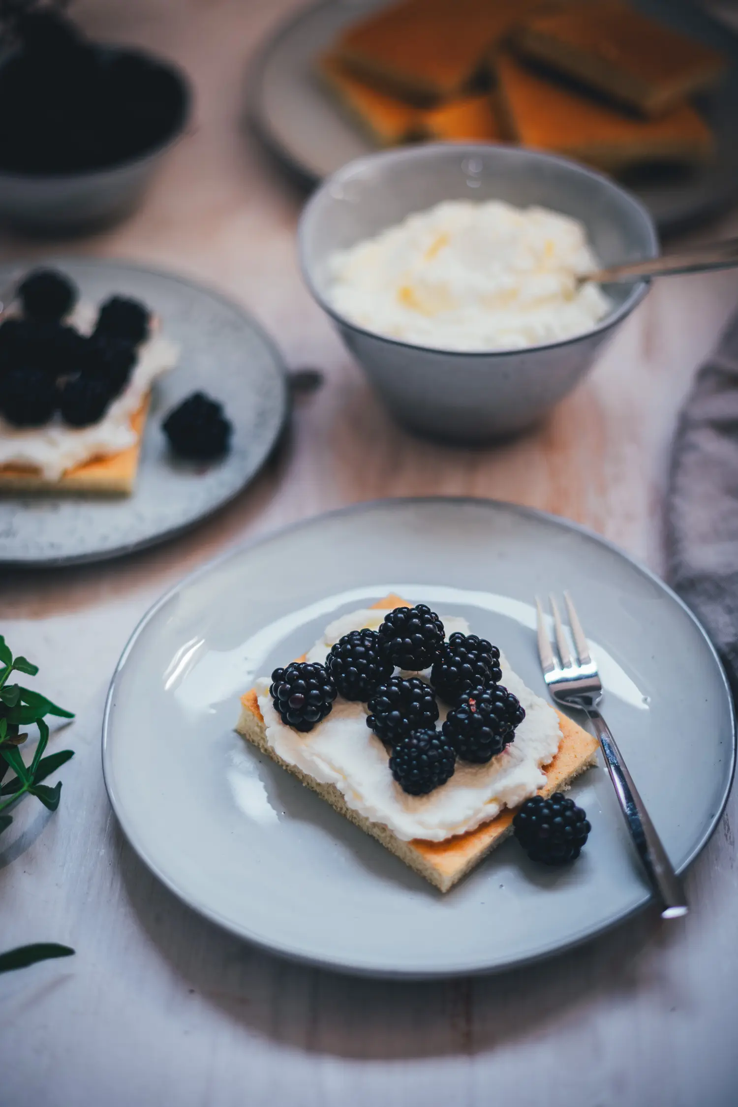 Ein Stück Hot Milk Sponge Cake mit Mascarpone-Creme und Brombeeren auf einem grauen Teller, dahinter eine Schale Creme.