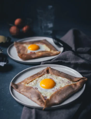 Seitliche Ansicht von zwei Tellern mit frisch gebackenen Galettes Complètes – französischen Buchweizenpfannkuchen aus der Bretagne, die mit Käse, Schinken und Spiegelei gefüllt sind.