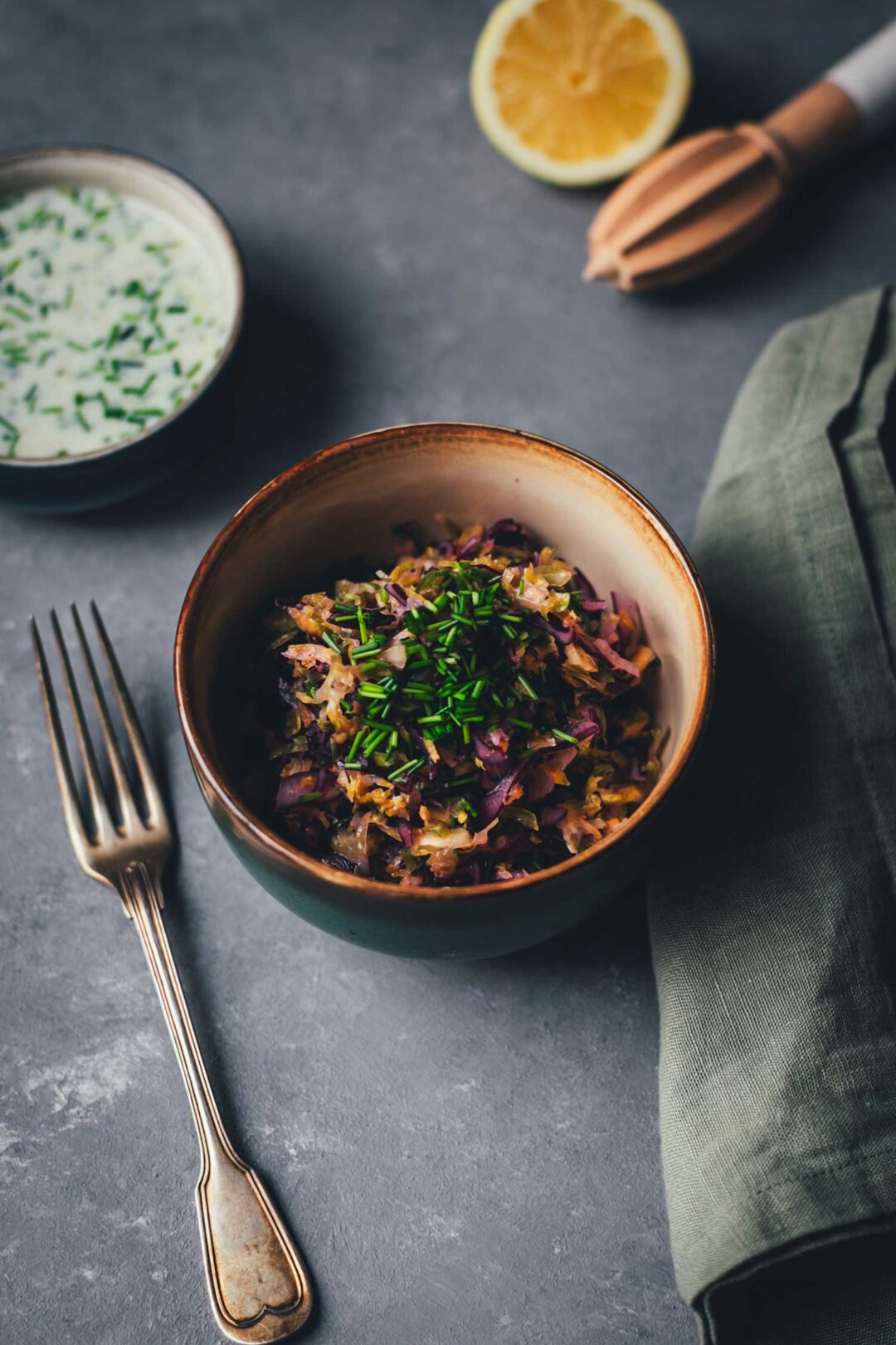 Krautsalat in Keramikschale mit Schnittlauch, daneben Gabel und Leinentuch – im Hintergrund Zitrone und Buttermilch-Zitronen-Dressing.