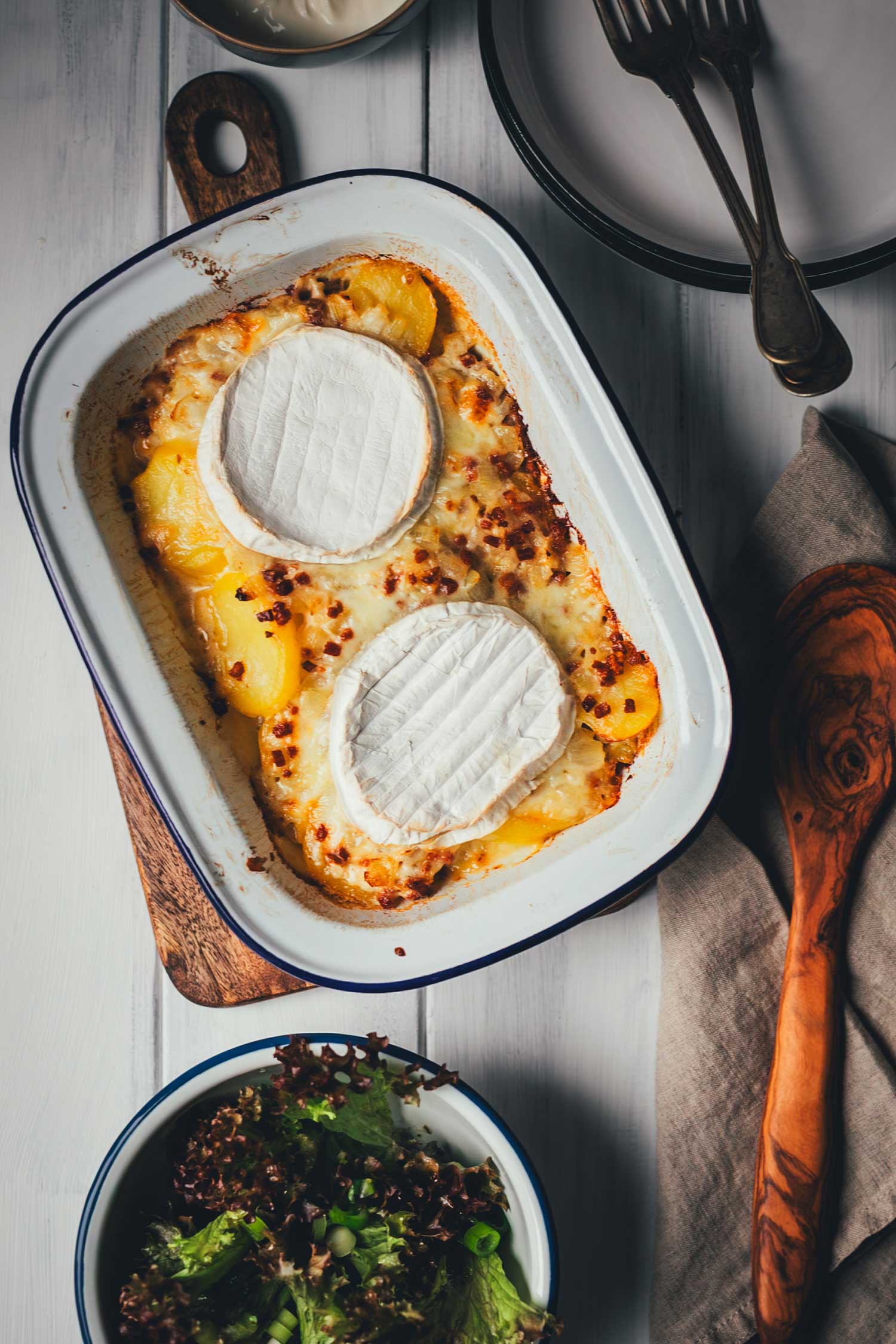 Servierfertige Tartiflette mit Camembert in der Auflaufform aus der Vogelperspektive, daneben ein frischer grüner Salat und Besteck auf einem hellen Holztisch.