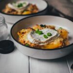 Seitliche Detailaufnahme einer Portion Tartiflette als quadratisches Bild: Der geschmolzene Camembert fließt über die knusprig gebräunten Kartoffelscheiben und krossen Speckwürfel.