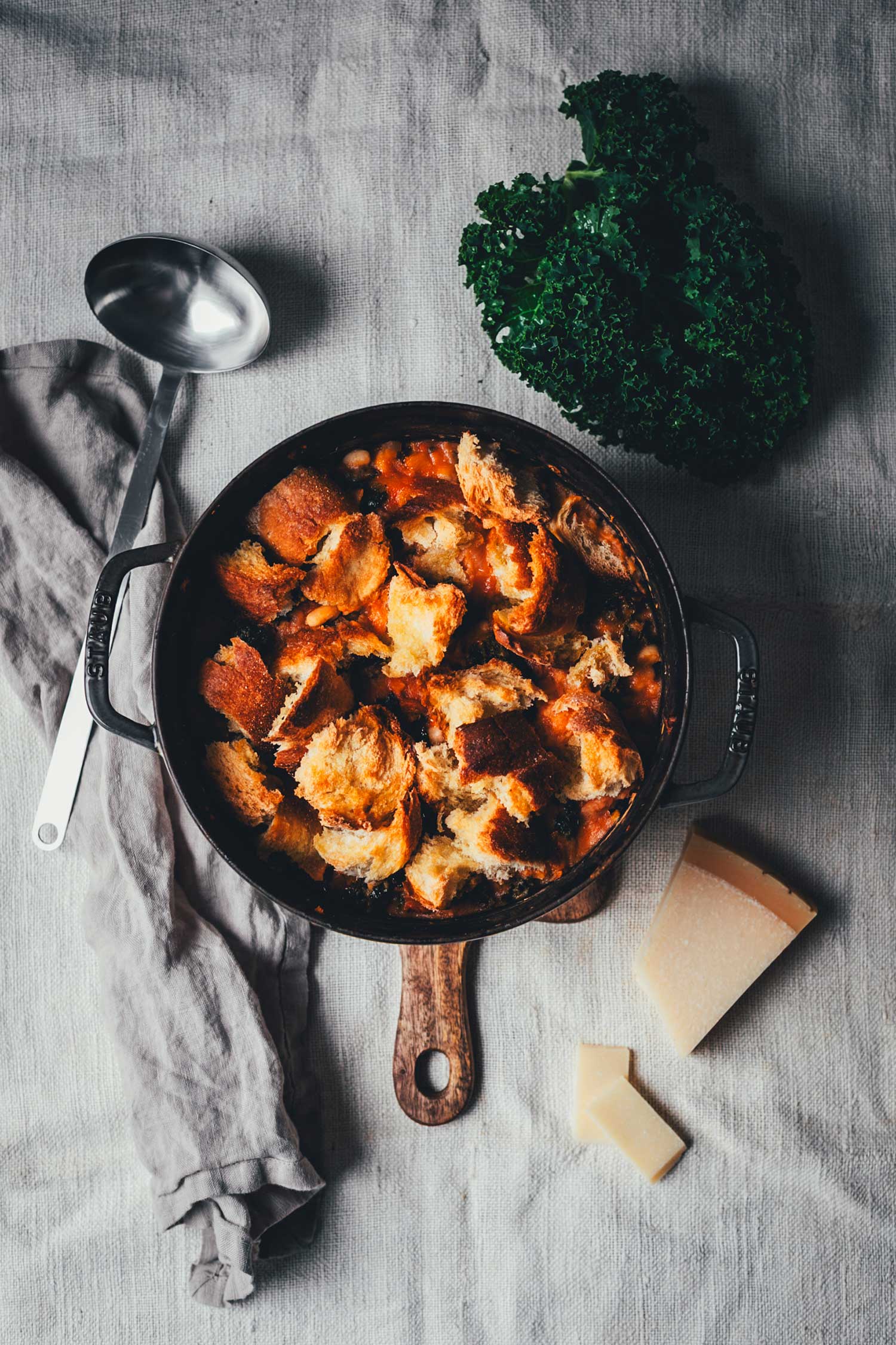 Topshot von fertig gekochtem und mit Brot getopptem Eintopf in einer gusseisernen Form. Drum herum sieht man ein Tuch, eine Schöpfkelle, ein Stück Parmesan und etwas Grünkohl. | moeyskitchen.com