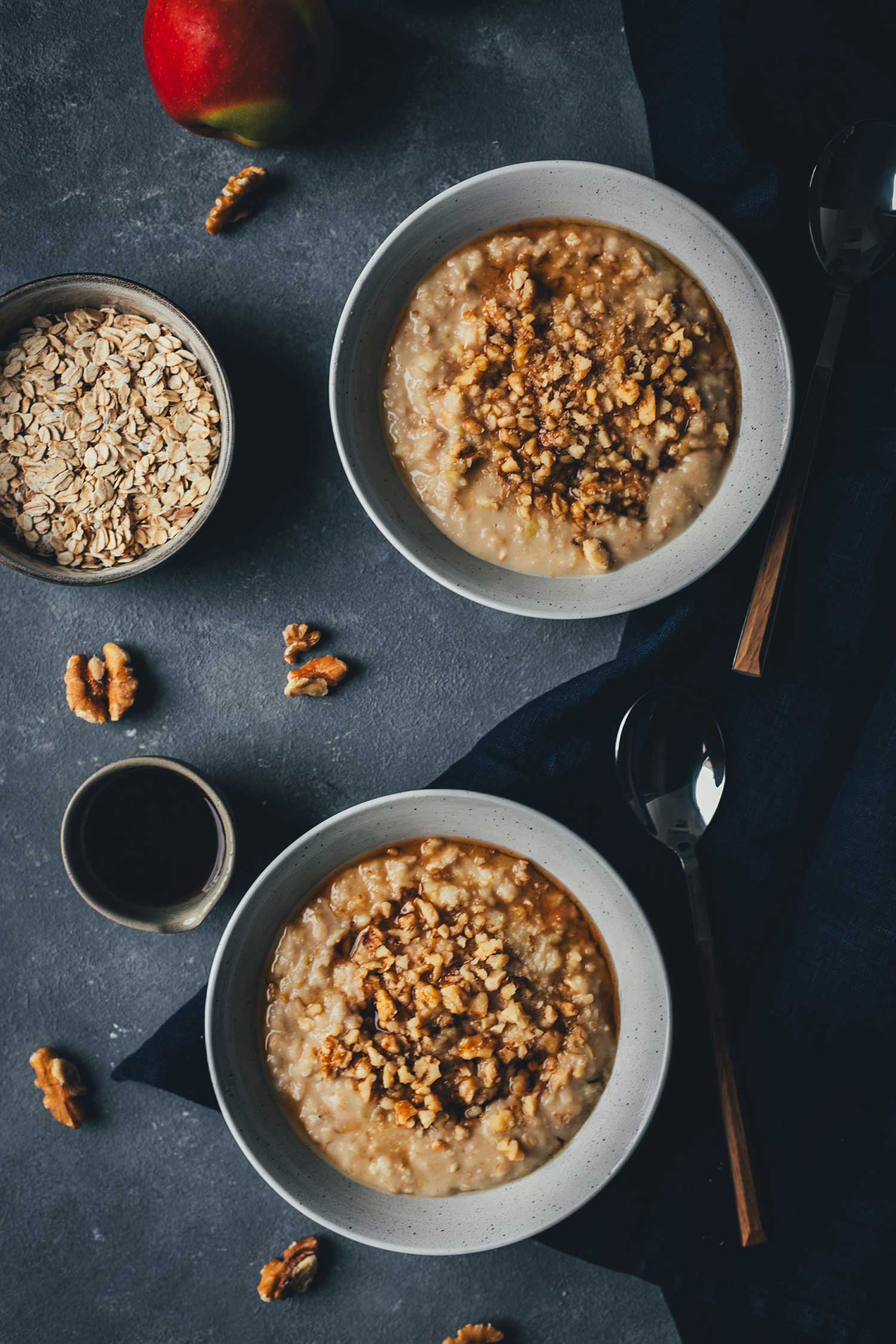 Topshot-Aufnahme von Apfel-Porridge in zwei Schalen. Daneben eine Schale mit Haferflocken, etwas Ahornsirup, Walnusskerne und Äpfel drumherum. | moeyskitchen.com