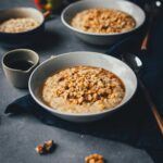 Quadratisches Bild von zwei Schüsseln mit cremigem Apfel-Porridge, seitlich fotografiert. Daneben sieht man Walnüsse, Haferflocken und Ahornsirup. | moeyskitchen.com