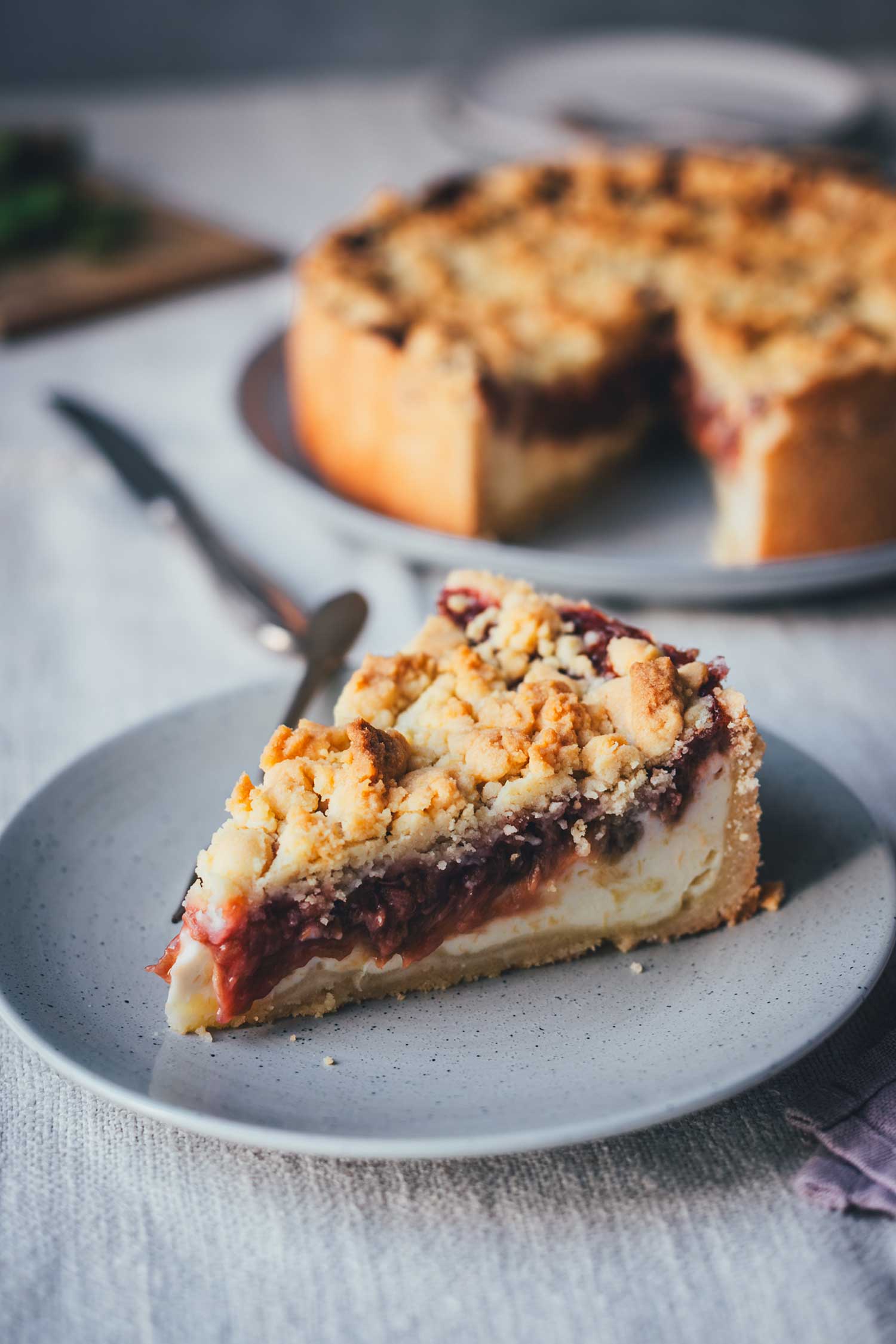 Stück Rhabarber-Käsekuchen mit Butterstreuseln und rotem Rhabarberkompott auf einem grauen Teller