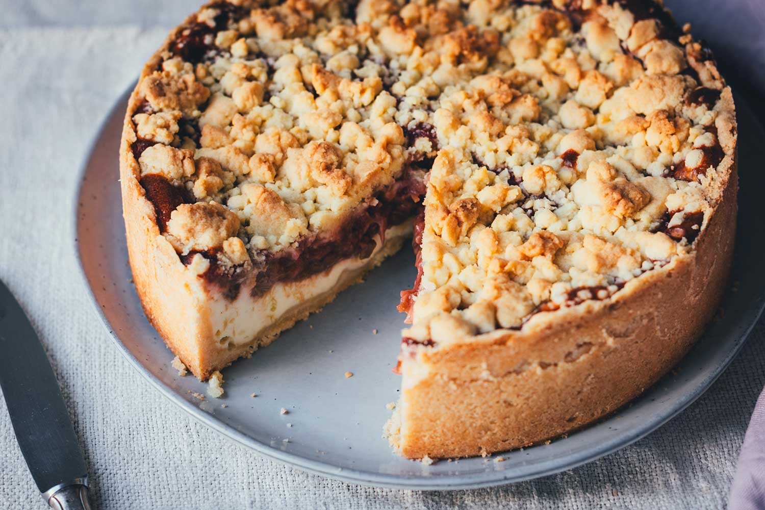 Rhabarber-Käsekuchen mit Streuseln auf einem Teller mit Messer, Draufsicht auf den angeschnittenen Kuchen, von dem schon ein Stück entnommen wurde.