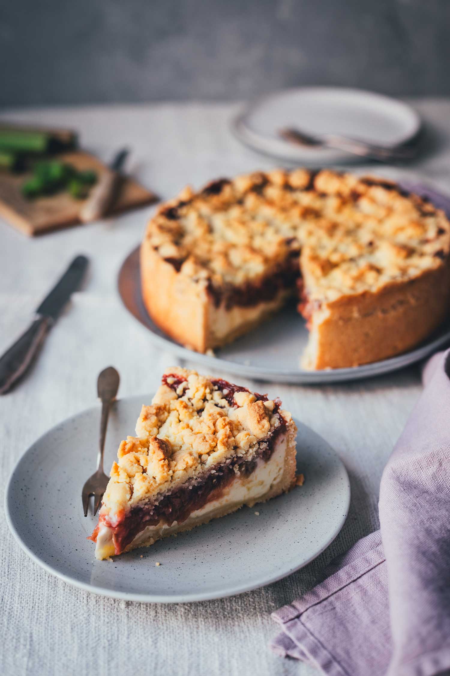 Ein Stück Rhabarber-Käsekuchen mit Streuseln auf einem Teller, ganzer Kuchen im Hintergrund