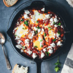 Topshot einer Pfanne mit Shakshuka aus dem Ofen. Eier in Tomaten-Paprika-Sauce geschmort und mit Feta und Petersilie bestreut serviert | moeyskitchen.com