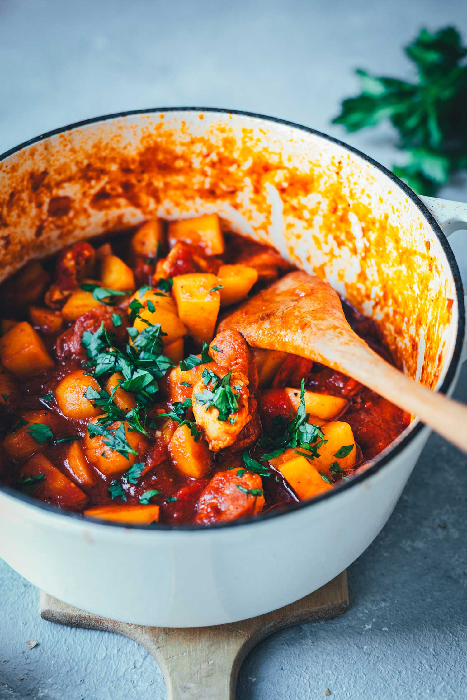 Spanischer Kartoffel-Paprika-Eintopf in einem gusseisernen Schmortopf auf einem Holzbrett serviert. Im Hintergrund Petersilie. | moeyskitchen.com