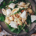 Topshot Nahaufnahme von einem Teller mit Muschelnudeln in einer Ziegenkäse-Sauce, serviert mit frischem Rucola und karamellisierten Walnüssen. Daneben liegt frisches Brot zum Servieren.