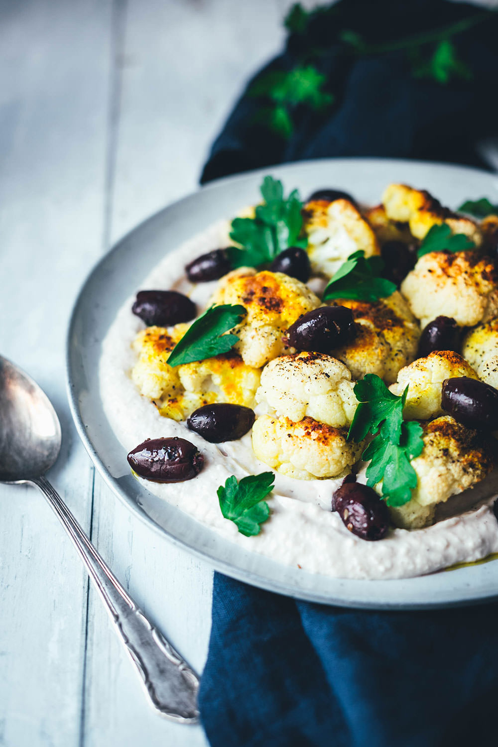 Rezept für gerösteten Blumenkohl aus dem Ofen. So gart der Kohl knusprig und dank asiatisch-orientalischer Gewürze auch lecker würzig! Serviert wird der Blumenkohl auf einer Bohnen-Creme aus weißen Bohnen, die mit griechischem Joghurt, Knoblauch und Zitronensaft verfeinert wird. Dazu gibt es gehackte Petersilie und leckere Kalamata-Oliven. Eine vegetarische Vorspeise oder Mezze, die lauwarm oder bei Raumtemperatur schmeckt. | moeyskitchen.com