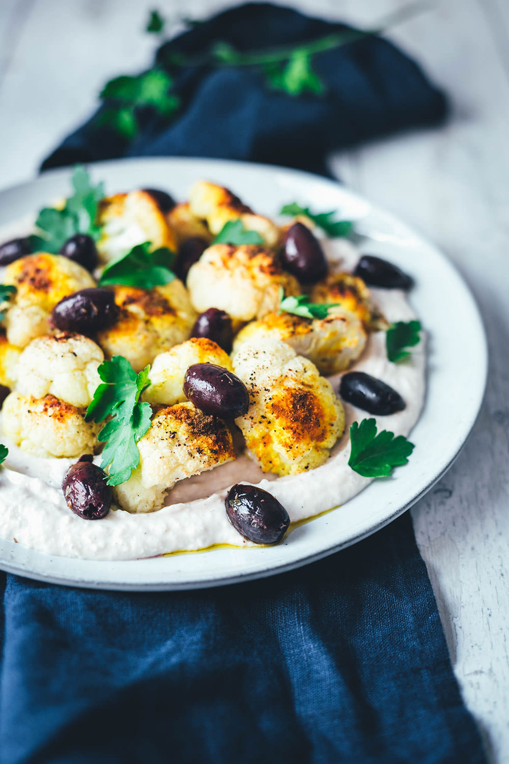 Rezept für gerösteten Blumenkohl aus dem Ofen. So gart der Kohl knusprig und dank asiatisch-orientalischer Gewürze auch lecker würzig! Serviert wird der Blumenkohl auf einer Bohnen-Creme aus weißen Bohnen, die mit griechischem Joghurt, Knoblauch und Zitronensaft verfeinert wird. Dazu gibt es gehackte Petersilie und leckere Kalamata-Oliven. Eine vegetarische Vorspeise oder Mezze, die lauwarm oder bei Raumtemperatur schmeckt. | moeyskitchen.com