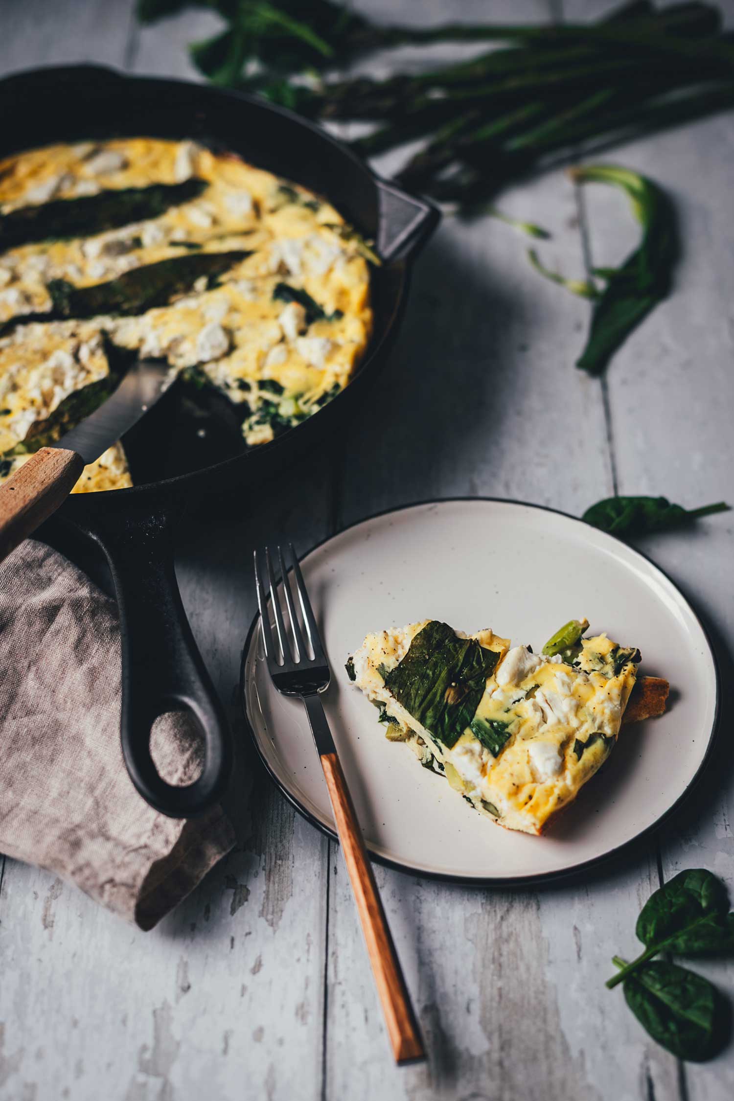 Ein Stück serviertbereite Frittata mit Bärlauch, Spinat, Spargel und Feta auf einem Teller. Im HIntergrund sieht man die Pfanne.