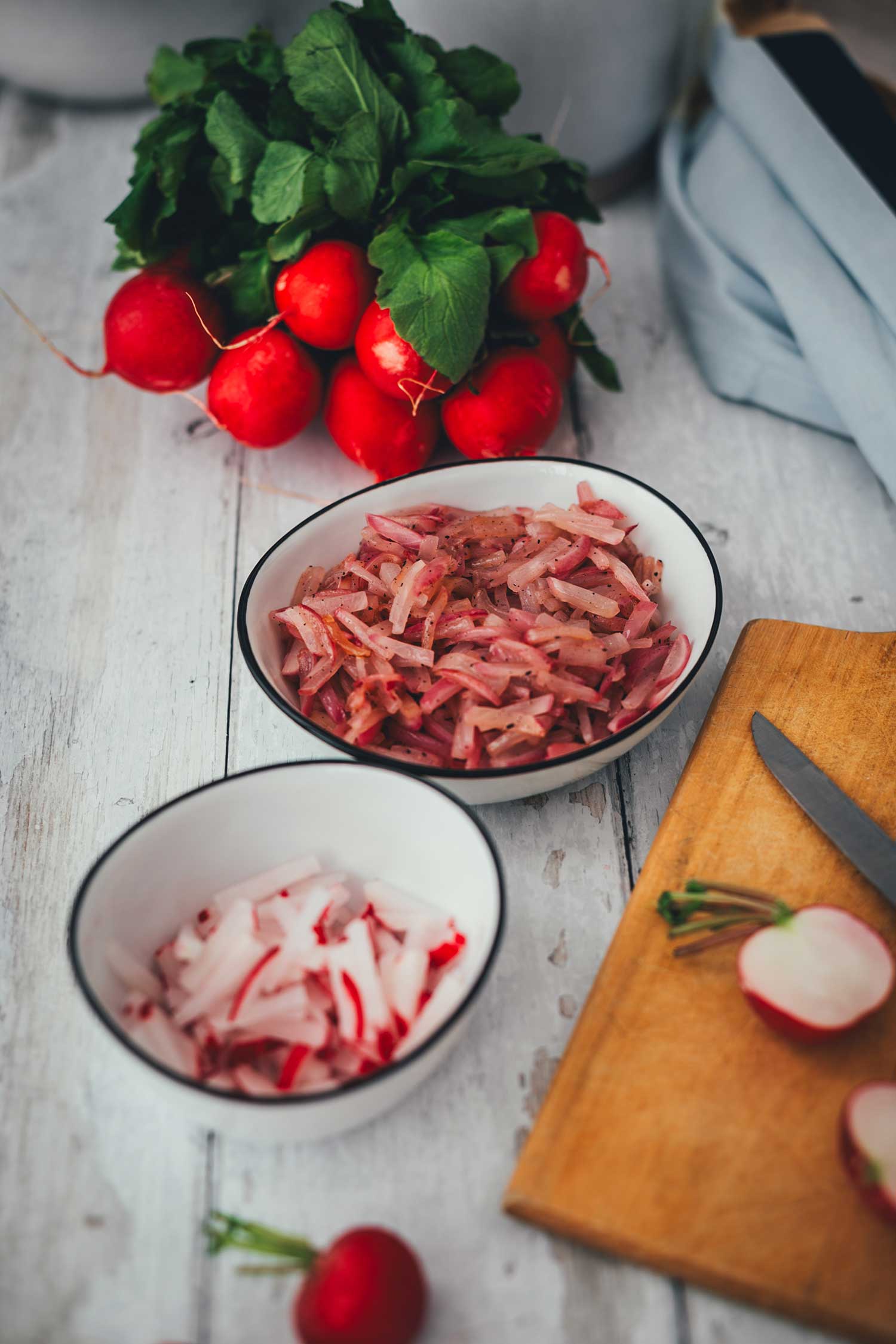 Vorbereitung von Radieschen für ein Obatzda Rezept: Ein Bund frische Radieschen, eine Schale mit rohen Radieschenstiften und eine Schale mit bereits sanft in der Pfanne gedünsteten Radieschen auf einem hellen Holzbrett.