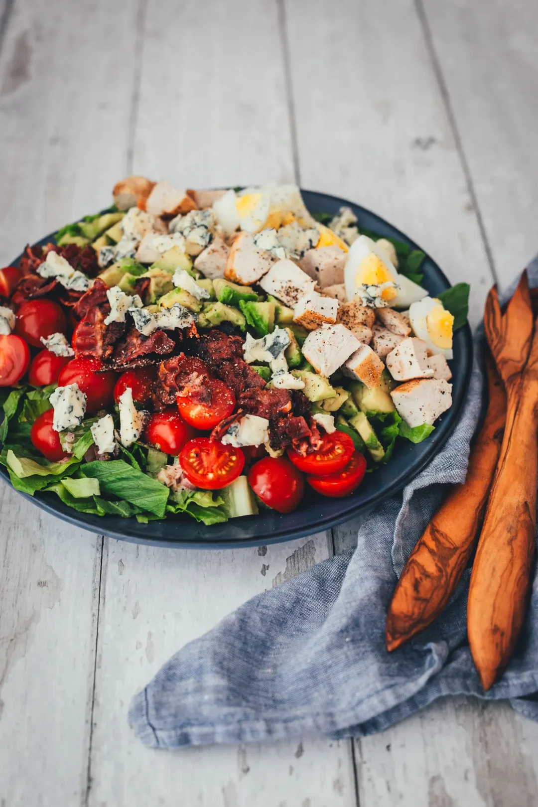 Angerichteter Cobb Salad auf dunklem Teller mit gebratener Hähnchenbrust, knusprigem Bacon, Ei, Avocado, Cocktailtomaten und Blauschimmelkäse auf Romana-Salat.