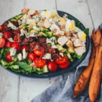 Angerichteter Cobb Salad auf dunklem Teller mit gebratener Hähnchenbrust, knusprigem Bacon, Ei, Avocado, Cocktailtomaten und Blauschimmelkäse auf Romana-Salat.