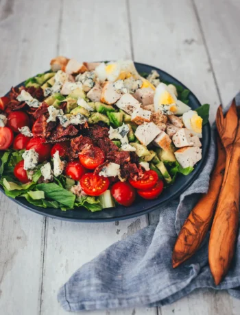 Angerichteter Cobb Salad auf dunklem Teller mit gebratener Hähnchenbrust, knusprigem Bacon, Ei, Avocado, Cocktailtomaten und Blauschimmelkäse auf Romana-Salat.