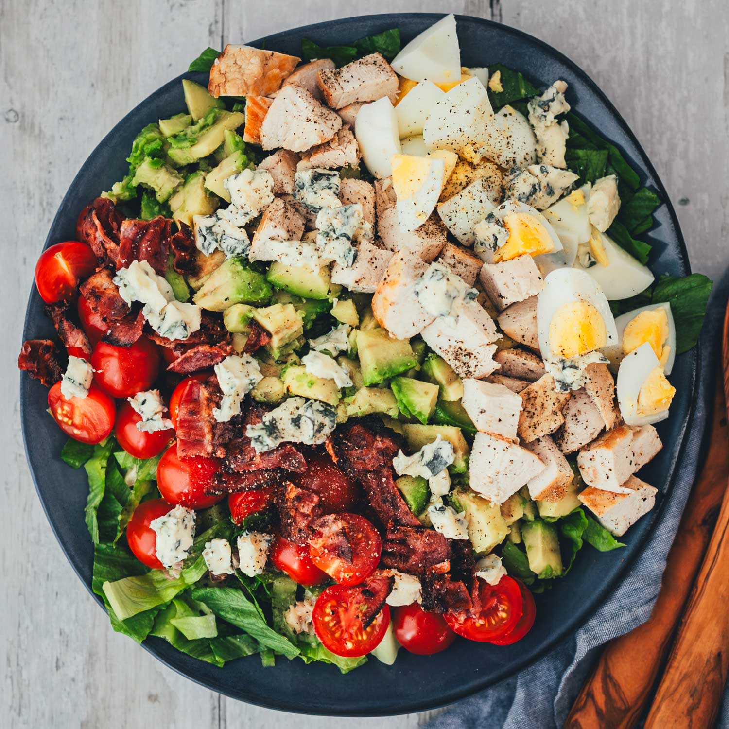 Cobb Salad von oben fotografiert, mit Hähnchen, Bacon, Ei, Avocado, Tomaten und Blauschimmelkäse in Reihen auf dunklem Teller angerichtet.