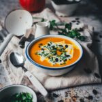 Leicht schräge Aufnahme einer Schüssel mit leuchtend oranger Kürbissuppe, mit Sahne und Kürbiskernen serviert. Darum Kräuter, Serviette, Löffel und Schöpfkelle, im Hintergrund ein Hokkaido-Kürbis. | moeyskitchen.com