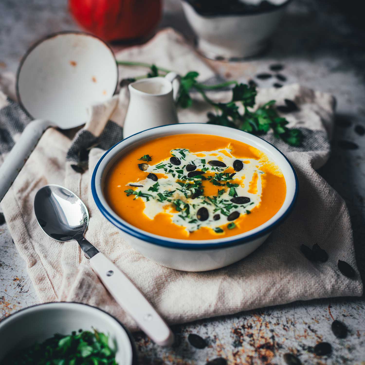 Leicht schräge Aufnahme einer Schüssel mit leuchtend oranger Kürbissuppe, mit Sahne und Kürbiskernen serviert. Darum Kräuter, Serviette, Löffel und Schöpfkelle, im Hintergrund ein Hokkaido-Kürbis. | moeyskitchen.com