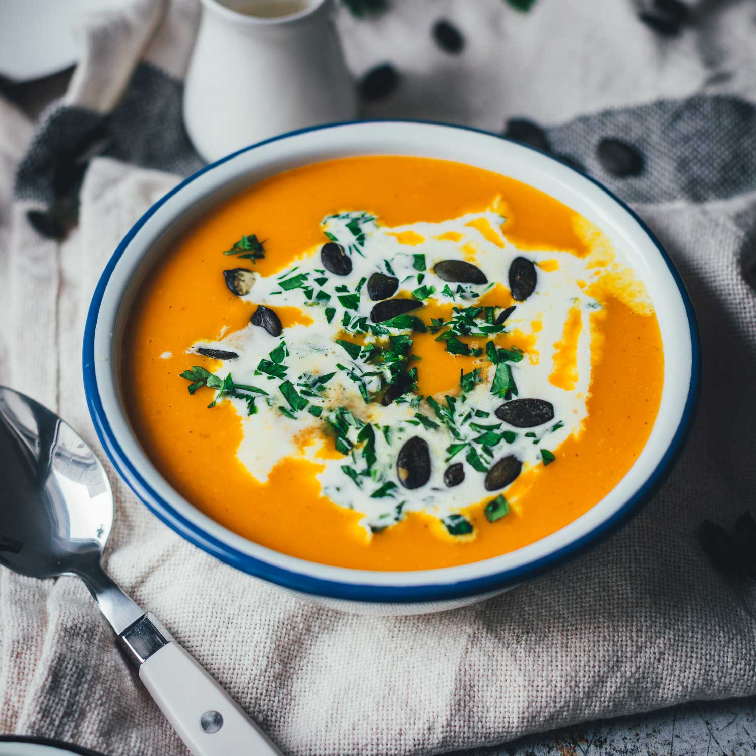 Servierbereite Kürbissuppe in einer weißen Emailleschüssel mit blauem Rand. Die klassische Kürbissuppe ist mit Schmand, Petersilie und Kürbiskernen getoppt.