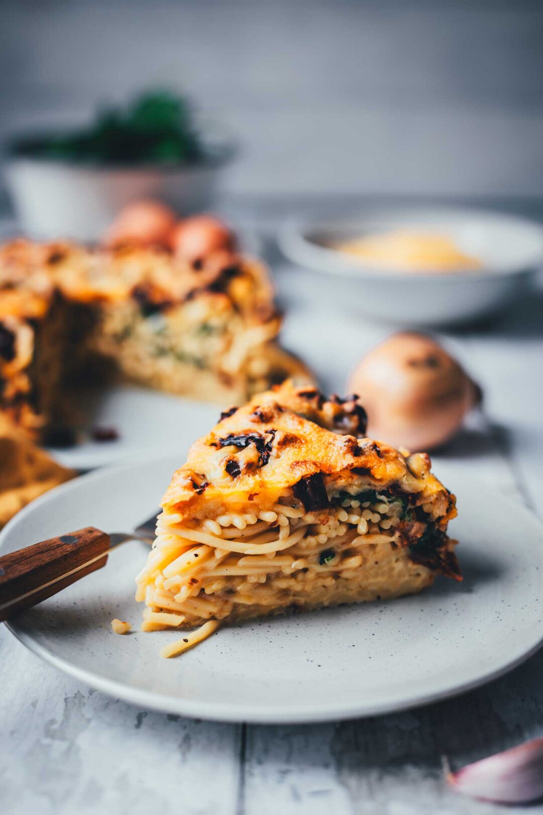 Auf einem Teller liegt ein Stück Spaghetti-Kuchen. Das ist ein Nudelauflauf aus Spaghetti, Spinat und getrockneten Tomaten, der in einer Springform gebacken und wie in ein Stück Kuchen geschnitten serviert wird. Man sieht die cremigen Schichten aus allen Zutaten und den ganzen Kuchen im Hintergrund.