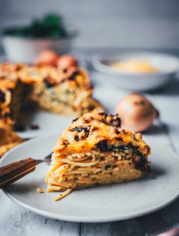 Auf einem Teller liegt ein Stück Spaghetti-Kuchen. Das ist ein Nudelauflauf aus Spaghetti, Spinat und getrockneten Tomaten, der in einer Springform gebacken und wie in ein Stück Kuchen geschnitten serviert wird. Man sieht die cremigen Schichten aus allen Zutaten und den ganzen Kuchen im Hintergrund.
