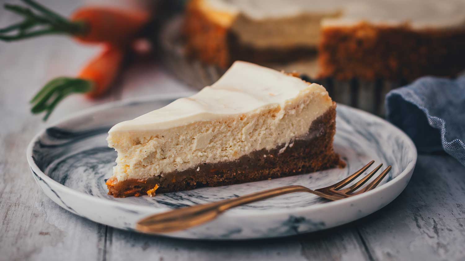 Nahaufnahme von einem Stück des Carrot Cake Cheesecakes auf einem marmomierten Teller. Man sieht sehr gut die Schichten aus Rüblikuchen als Boden und dem typischen Cheesecake-Topping. Im HIntergrund steht der angeschnittene Kuchen, daneben liegen ein paar Möhren.
