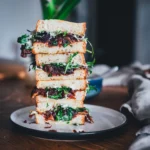 Seitliche Ansicht von vier übereinandergestapelten Sandwiches: Hummus-Sandwich mit Bohnen-Hummus und karamellisierten Zwiebeln, serviert auf einem weißen Teller auf einem Holztisch.
