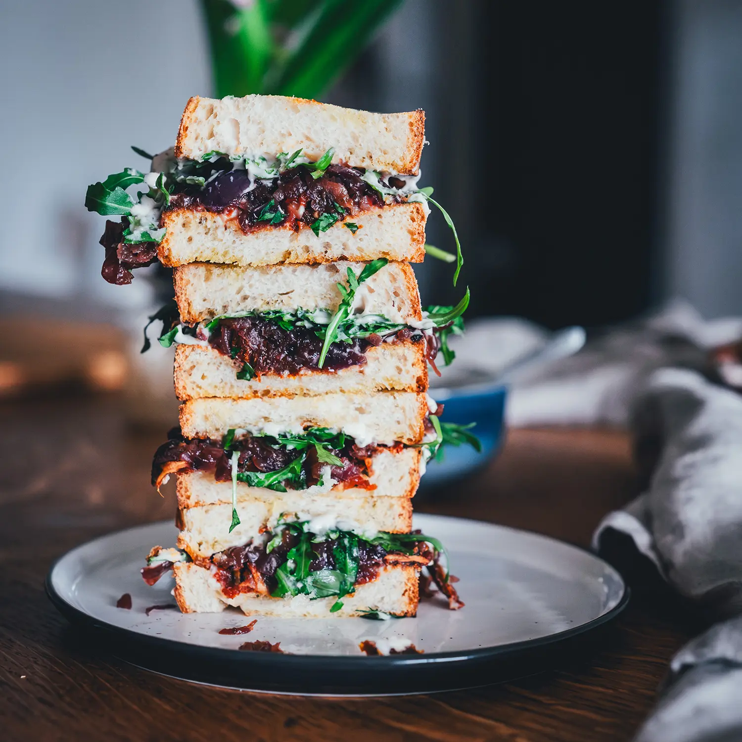 Seitliche Ansicht von vier übereinandergestapelten Sandwiches: Hummus-Sandwich mit Bohnen-Hummus und karamellisierten Zwiebeln, serviert auf einem weißen Teller auf einem Holztisch.