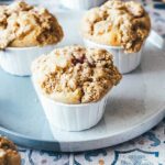 Frisch gebackene Erdbeer-Rhabarber-Muffins mit knusprigen Butterstreuseln stehen auf einem Teller. Im Hintergrund liegen frische Erdbeeren und Rhabarber.